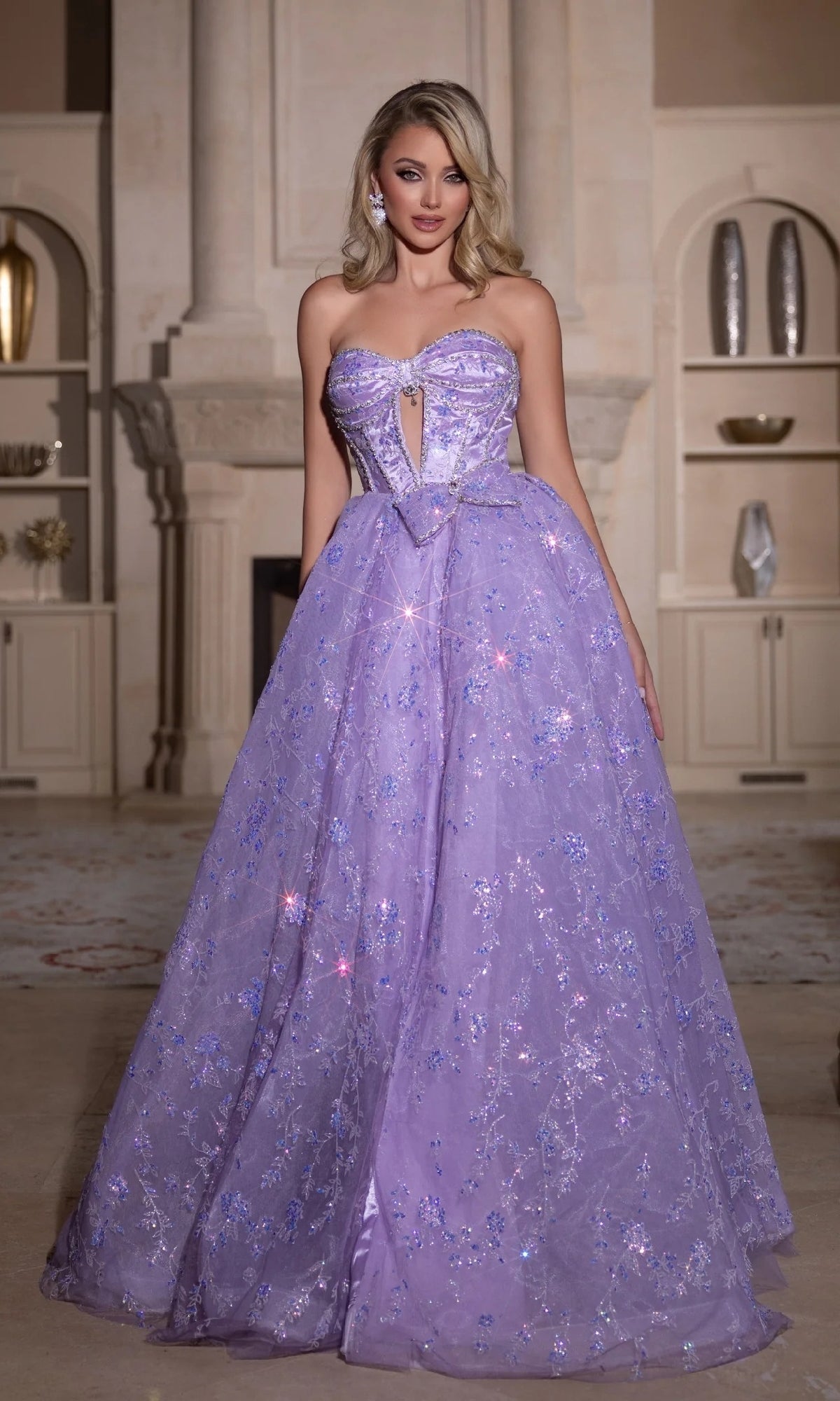 A woman wears the Portia and Scarlett PS25315 long prom dress with sparkling embellishments, standing indoors by built-in shelves and a fireplace.